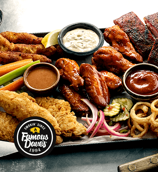 Famous Dave's BBQ Platter on a black tray with roasted chicken, chicken wings, breaded chicken, ribs, pickles, shoestring pickles, pickled onions, celery and carrot sticks, and various dipping sauces