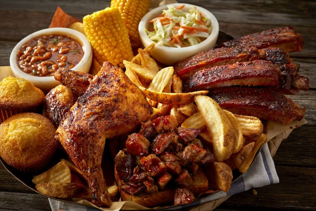 Image of Famous Dave's All-American BBQ Feast® with chicken legs, ribs, corn on the cob, cornbread, wedge fries, baked beans, and cole slaw all stacked on top of each other