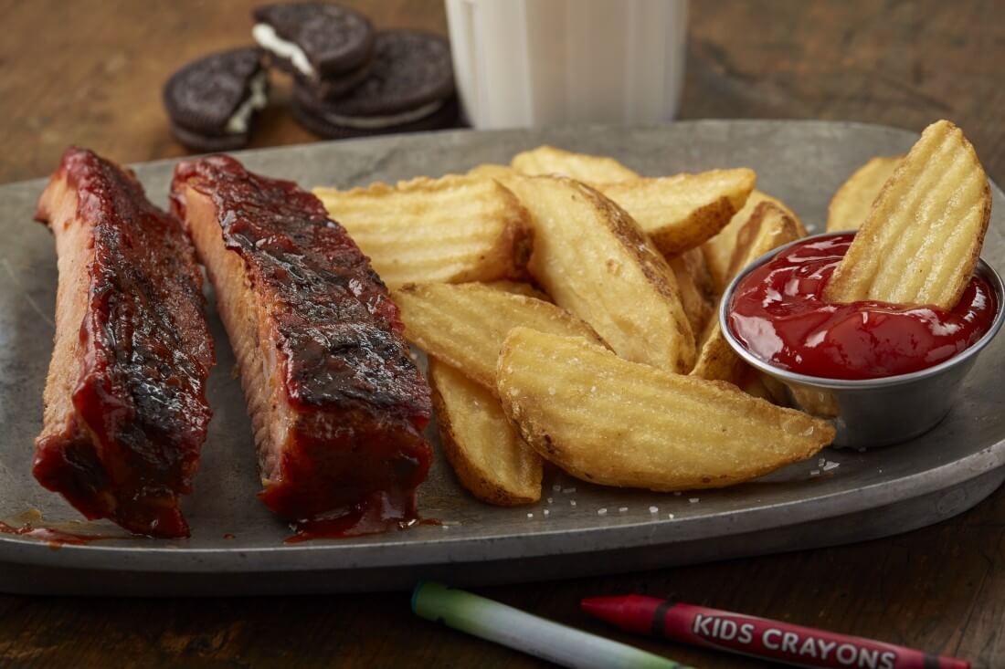 Two individual ribs smothered in BBQ sauce, a few wedge fries, and a small ramekin of ketchup on a gray plate