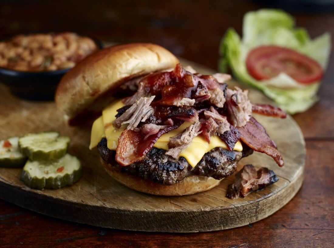 Image of the Ultimate Burger, topped with chopped pork and bacon, served on a plank of wood, with a bowl of baked beans on the side, pickles, lettuce and tomatoes on the side