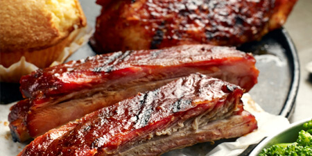Two individual ribs covered in BBQ sauce in the foreground, behind the ribs is a piece of cornbread and BBQ chicken.
